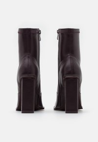 Botas de tobillo moradas oscuras con tacones cuadrados y cremalleras laterales, vistas desde atrás sobre un fondo blanco liso.