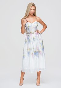 Femme blonde portant une robe midi blanche à fleurs avec des fines bretelles et des sandales argentées à talons hauts, debout devant un fond clair uni.