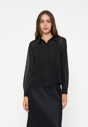 Femme portant une blouse noire à manches longues transparentes et une jupe noire, debout devant un fond clair uni.