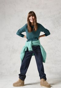 Pull en teal texturé superposé sur un haut teal clair, associé à un jean bleu marine et des bottines marron. Modèle souriant sur un fond clair.