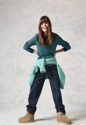 Türkisfarbener, strukturierter Pullover, über einem hellturkisen Oberteil getragen, kombiniert mit dunkelblauen Jeans und braunen Stiefeletten. Model lächelt vor einem hellen Hintergrund.