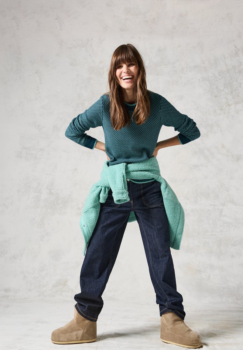 Pull en teal texturé superposé sur un haut teal clair, associé à un jean bleu marine et des bottines marron. Modèle souriant sur un fond clair.