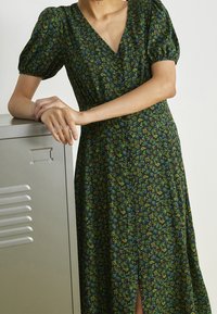 Robe maxi fleurie avec un fond sombre, dotée de manches courtes bouffantes, d'un décolleté en V et d'une fente à l'avant. Fabriquée en tissu léger avec de petits motifs.