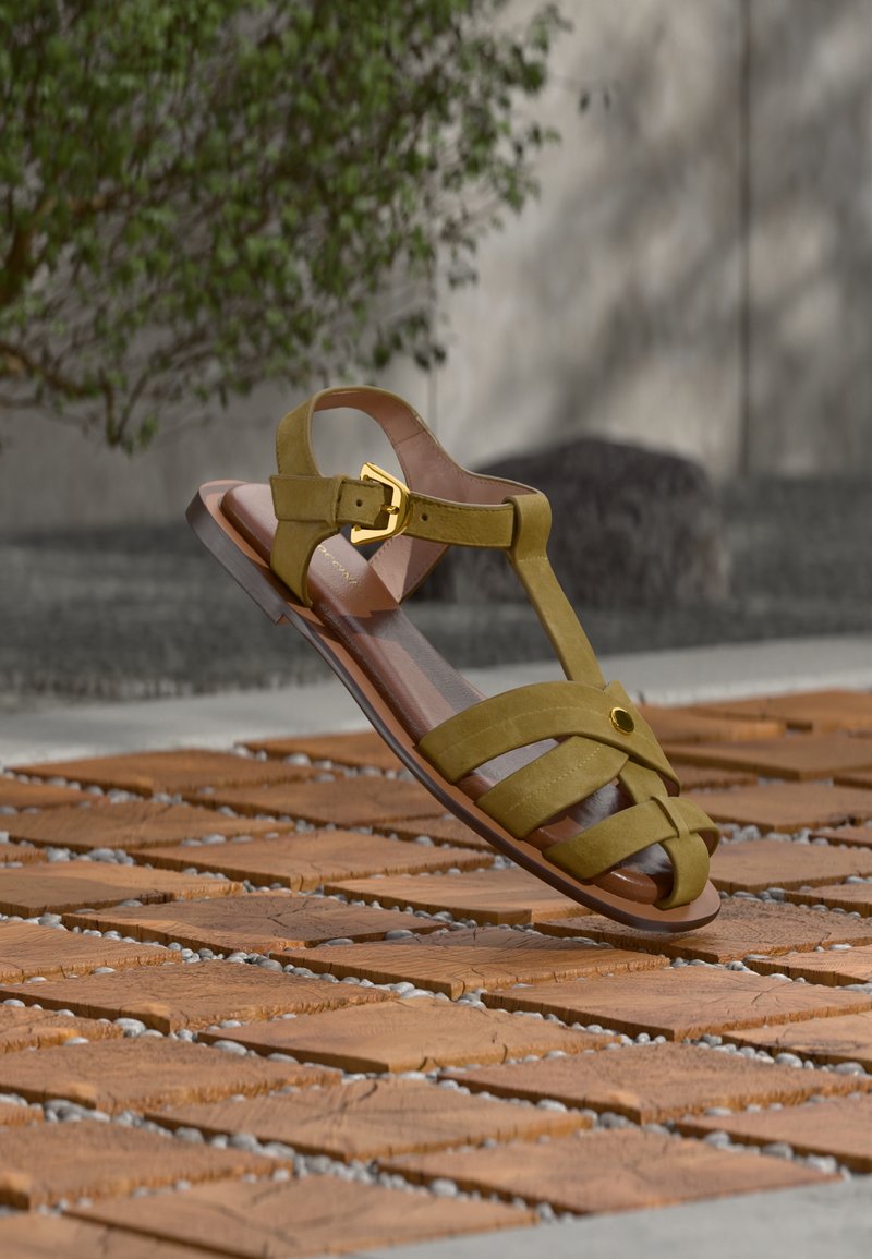 Δερμάτινο σανδάλι σε ελιά πράσινο με χρυσαφί αγκράφα και λουριά, εμφανίζεται να αιωρείται πάνω από υφασμένους ξύλινους πλακιδίους σε υπαίθριο περιβάλλον.