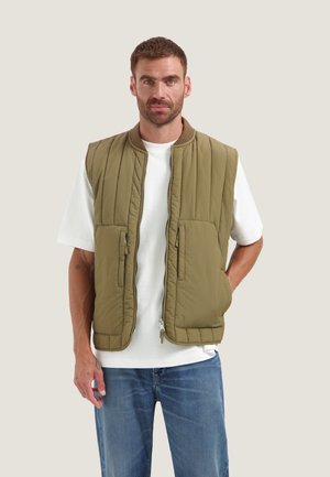 Uomo con gilet imbottito senza maniche verde oliva sopra una camicia bianca a maniche corte e jeans blu, in piedi con una mano in tasca.