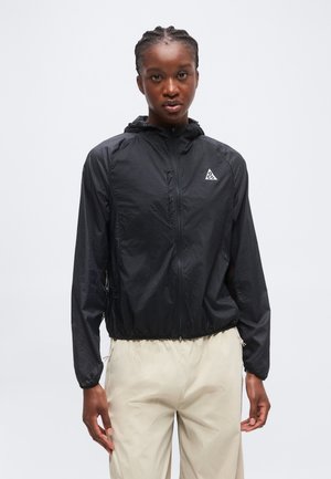 Person wearing a black hooded jacket with a white logo on the chest and light beige pants against a plain light background.