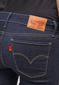 Jeans Levi's de color azul oscuro con costuras amarillas distintivas, bolsillo trasero y parche de cuero color marrón claro de la marca justo encima de la cinturilla, en una persona que lleva una camiseta negra.