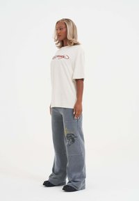 Lys beige bomulds t-shirt med rød grafisk tekst, parret med løse grå denim bukser med gule og sorte broderede detaljer.