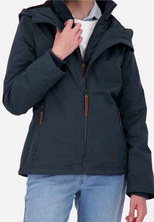 Personne portant une veste zippée bleu marine avec des tirettes de fermeture éclair marron et un jean bleu clair, ajustant le col de la veste d'une main.