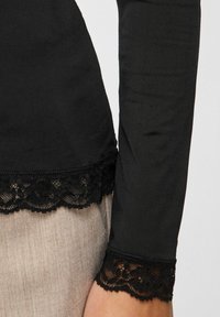 Svart langermet topp med blondekant ved ermekanter og nederst på plagget. Myk tekstur og tettsittende design, vist med et lyst stoff under.
