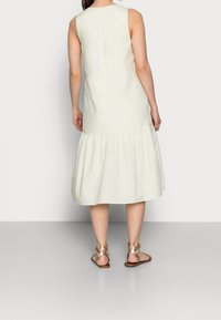 Hellgrünes, ärmelloses Kleid mit einem gestuften Saum, aus weichem Material gefertigt. Das Model trägt metallische Sandalen und zeigt freiliegende Beine.