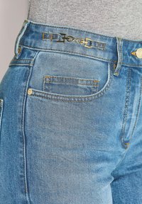 Nahaufnahme von hellblauen Jeans mit goldenen Beschlägen und Nähten, getragen mit einem grauen Oberteil.