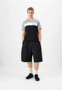 Ung mand står med hænderne i lommerne iført en grå og sort Rip Curl T-shirt, sorte brede shorts, hvide sokker og sorte sneakers.