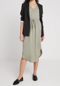 Czarna marynarka na jasnookiej, zielonej sukience do kolan z wiązaniem w talii. Sukienka ma dekolt w serek i boczne rozcięcia, zestawiona z czarnymi loafersami.