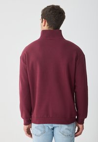 Burgunderfarget genser med høy krage, ribbestrikkede mansjetter og kant, laget av myk stoff. Synlige søm detaljer på skuldrene.