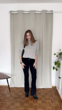 Maglione a maglia grigio chiaro; jeans neri a zampa d'elefante; stivaletti neri; sfondo texturizzato; pianta verde; pavimento in legno; tavolo semplice con gambe sottili.