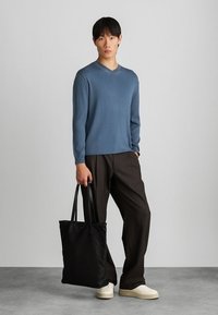 Μπλε ριμπ φανέλα μακρύ μανίκι με V-λαιμό, συνδυασμένη με χαλαρούς καφέ παντελόνες και μια μαύρη τσάντα tote, με λεία υφή και καθαρό σχεδιασμό.