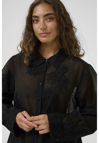 Black sheer blouse with button closure, detailed embroidery, and a classic collar. Features long sleeves and textured fabric.