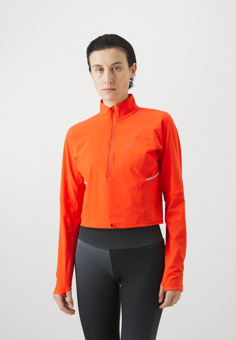 Lysoransje langermet topp med halv-glidelås krage, som har en slank passform og reflekterende detaljer på sidene. Kombinert med mørke leggings.
