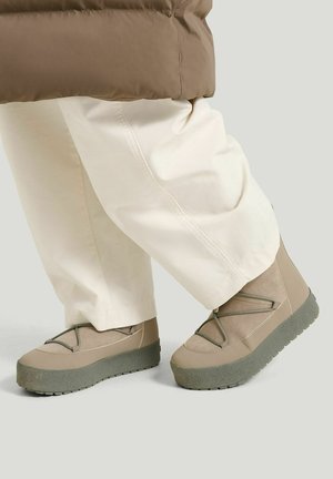 Person trägt beige Plateauschuhe mit dicken Sohlen und cremefarbene Hosen vor einfarbigem Hintergrund.