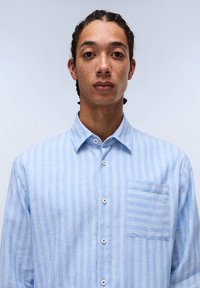 Camisa de botones a rayas azul claro con un bolsillo, hecha de una tela ligera que presenta rayas verticales y botones azules.