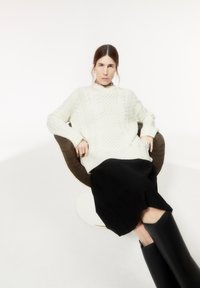 Witte oversized gebreide trui met textuurpatronen, gecombineerd met een zwarte rok en zwarte kniehoge laarzen, zittend op een bruine stoel.