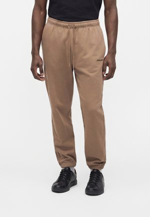Mand iført brune joggingbukser med elastiske manchetter og sorte sneakers, stående mod en ensfarvet lys baggrund.