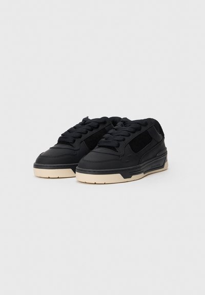 Ein Paar schwarze Low-Top-Sneaker mit dicken weißen Sohlen und schwarzen Schnürrsenkeln auf einem einfarbigen hellgrauen Hintergrund.