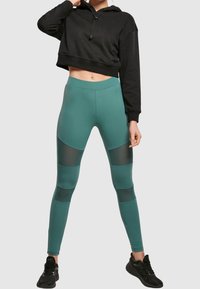 Svart kort hoodie med dragsko. Teal leggings med nätpaneler i en geometrisk design. Svarta träningsskor fulländar outfiten.