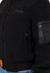 Giacca in pile nera con superficie texturizzata, tasca con zip in tessuto nero sulla manica e etichetta arancione sull'orlo.