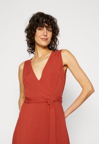 Vestido envolvente vermelho com um decote em V profundo, design sem mangas e detalhe de cintura amarrada. Feito de um tecido leve, com uma textura suave.