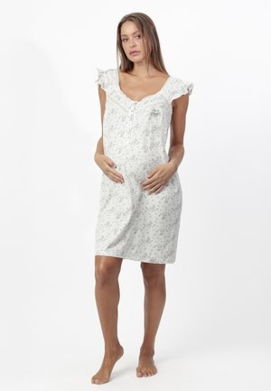 Mujer embarazada descalza, de pie, vestida con un vestido premamá ligero sin mangas con estampado floral y hombros con volantes, con las manos apoyadas en el vientre.