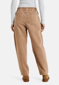 Pantalones de pierna ancha de color marrón claro con cintura elástica, tejido texturizado y dos bolsillos traseros. Se llevan con una camiseta de rayas de manga larga y botas.