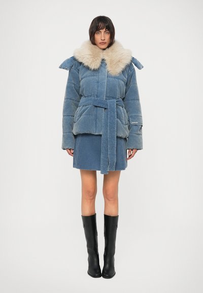 Vrouw in een blauw corduroy pufferjack met riem en een grote crèmekleurige bontkraag, bijpassende rok en zwarte kniehoge laarzen, rechtopstaand.