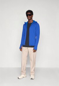 Muž v modrej mikine na zips, čiernom tričku, béžových nohaviciach, bielych teniskách a veľkých čiernych slnečných okuliaroch stojí proti jednému svetlému pozadiu.