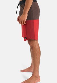 Shorts de bain rouges et marron en tissu extensible avec un design à rayure sur le côté, dotés d'une taille à cordon de serrage et d'un patch logo.