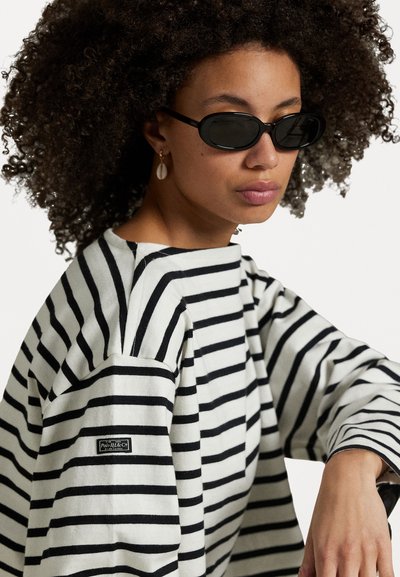 Chemise à rayures noires et blanches à manches longues, en coupe décontractée, dotée d'un col rond et d'un petit patch logo sur la manche. Lunettes de soleil ovales noires.