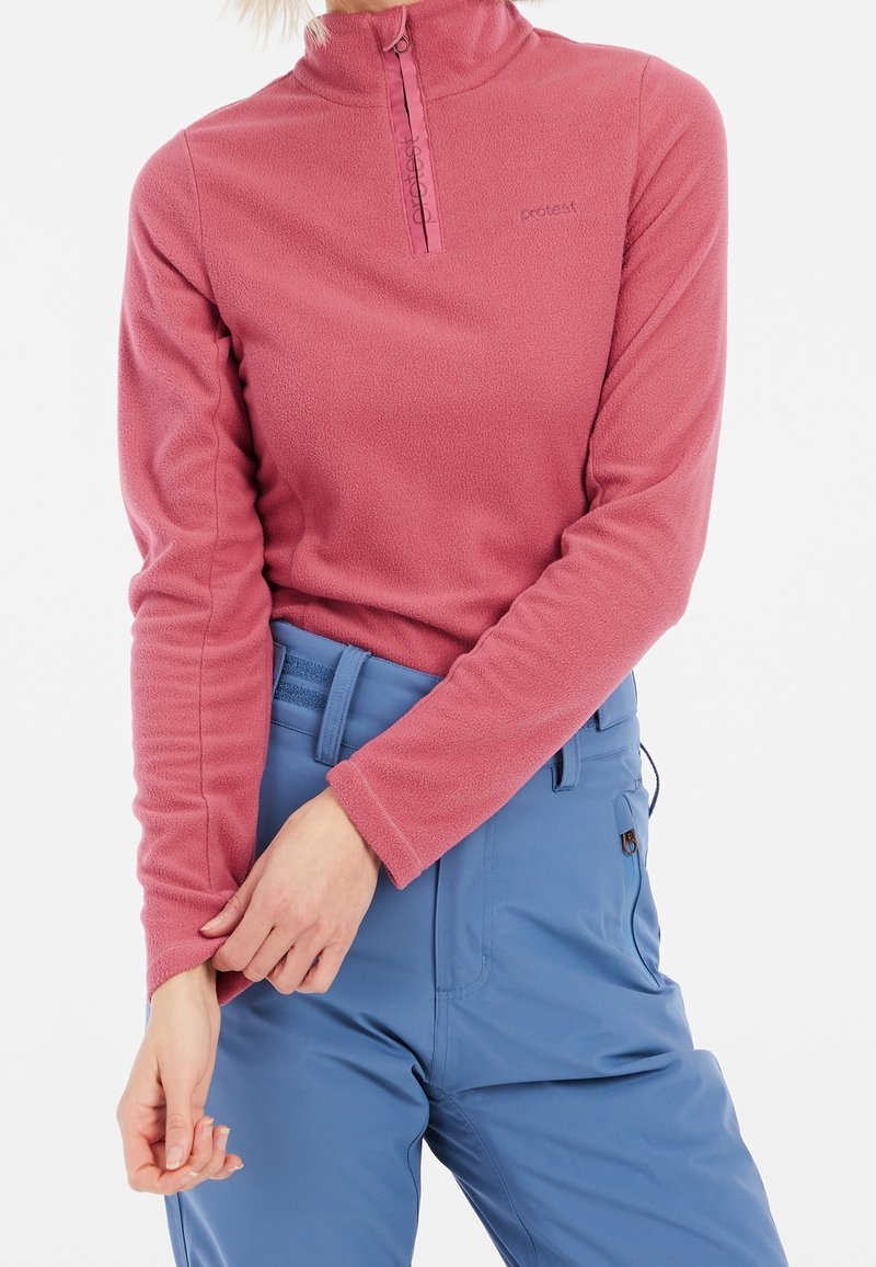 Femme portant un haut polaire rose à fermeture éclair quart et un pantalon bleu taille haute, ajustant la manche polaire avec la main.