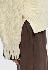 Gros plan sur la manche d'un pull en maille beige avec bordure noire portée sur un pantalon marron, montrant un détail d'ouverture latérale et une coupe décontractée.