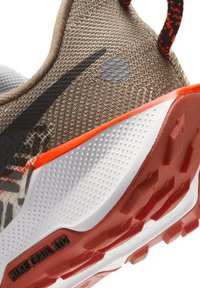 La zapatilla de trail de Nike presenta un upper texturizado en color marrón, acentos en negro y gris, una franja de color naranja brillante y una suela roja con un patrón para ofrecer tracción.