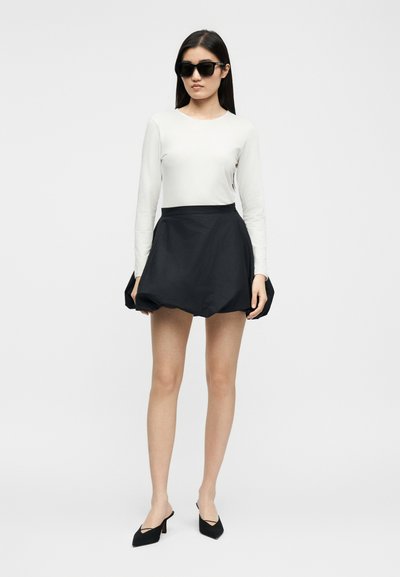 Femme portant des lunettes de soleil noires, un haut blanc à manches longues, une mini-jupe noire bouffante et des escarpins noirs à bouts pointus, debout sur un fond clair uni.