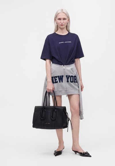 Femme aux longs cheveux clairs portant un t-shirt bleu marine Marc Jacobs, une jupe grise à fermeture éclair New York, des chaussures pointues noires, tenant un grand sac à main noir.