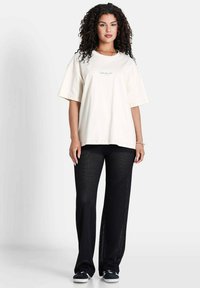 T-shirt en coton beige surdimensionné avec des manches courtes et un logo subtil ; associé à un pantalon en maille noire texturée, présentant une coupe décontractée et des fentes sur les côtés.