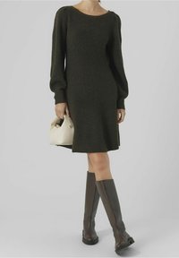 Robe en maille vert foncé avec manches longues bouffantes, encolure arrondie et fentes latérales. Associée à des bottes marron montant jusqu'au genou et un sac à main clair.