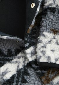 Veste en polaire avec un motif géométrique noir, blanc et gris, dotée d'une texture douce et d'une doublure noire, fermée par un bouton-pression en métal.