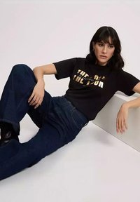 Czarny t-shirt z grafiką, na którym znajduje się złoty tekst, zestawiony z wysokim stanem ciemnych dżinsów, na neutralnym tle.