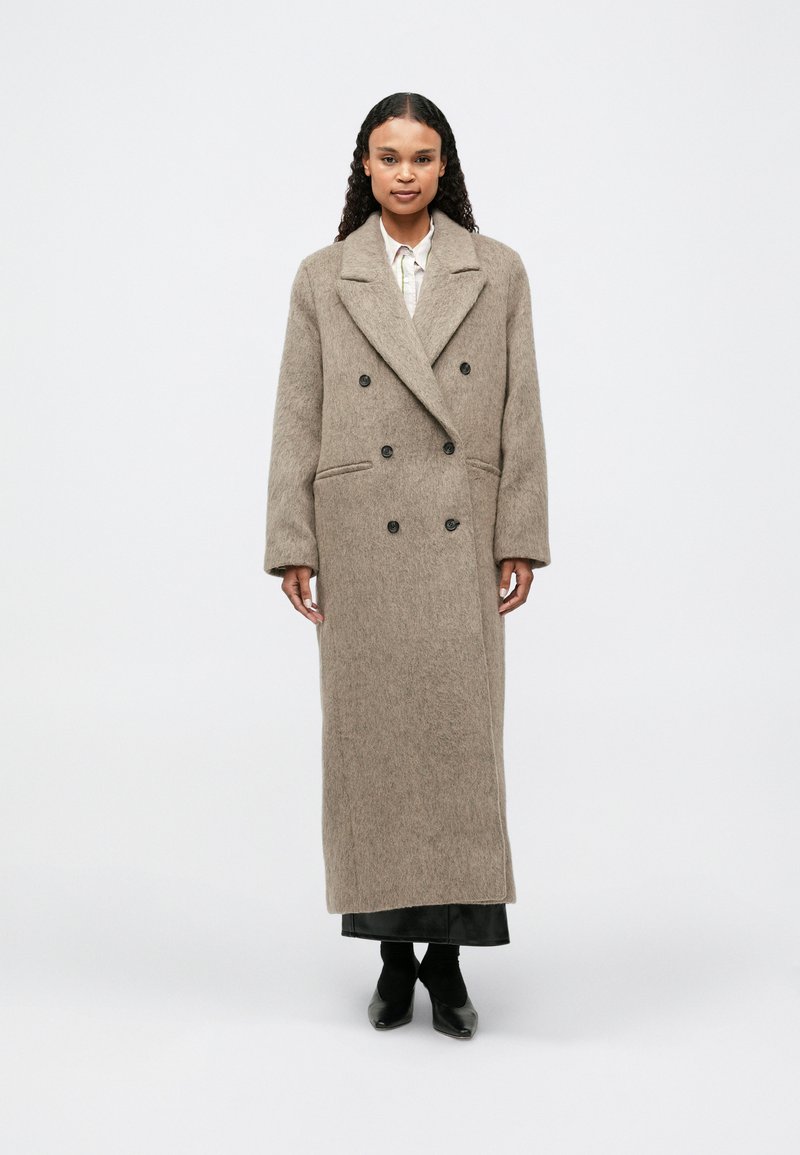 Długi, podwójnie zapinany płaszcz z miękkiej, beżowej mieszanki wełny. Posiada klapowy kołnierz, dwie przednie kieszenie i czarne guziki. Dopasowany, prosty krój.