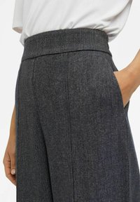 Pantalons gris texturés avec une large ceinture, poches latérales et coupe ajustée. Le matériau a un motif subtil, améliorant l'ensemble du design.
