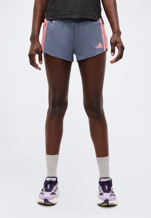 Atlet iført grå Adidas løbeshorts med koralstriber, sort top, grå sokker og lilla sportssko på ensfarvet baggrund.