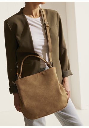 Frau trägt olivgrünen Blazer und weißes Hemd, hält eine große beige Ledertasche mit Schultergurt und Griff.